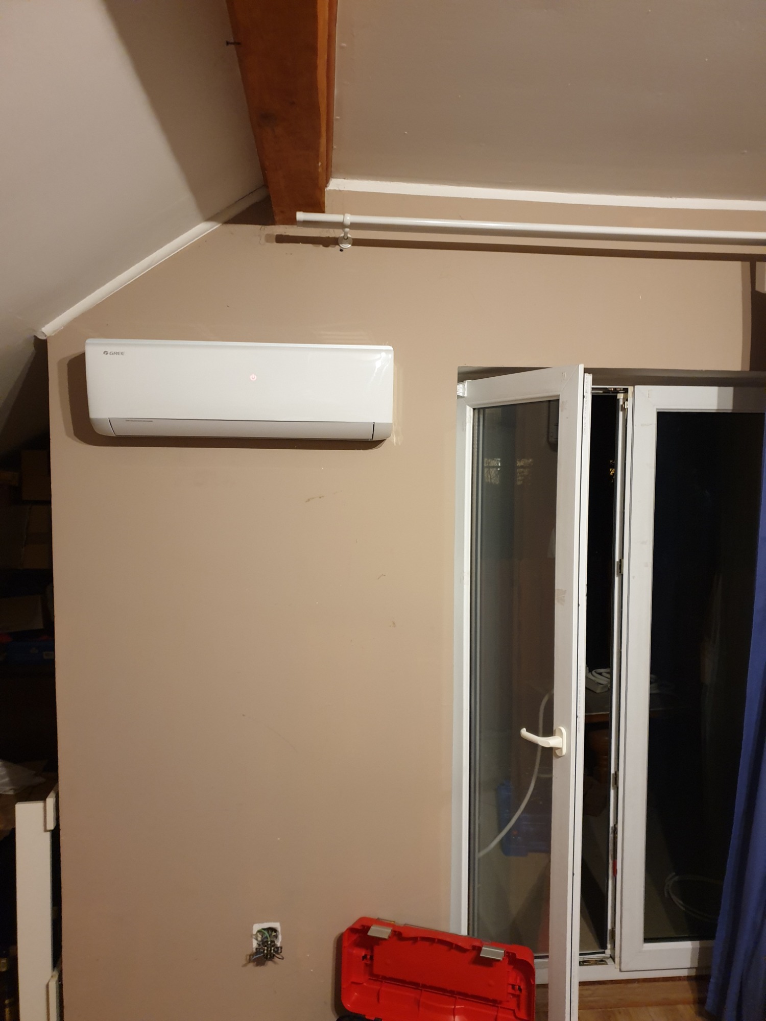 Biały klimatyzator marki Gree zamontowany na beżowej ścianie pod skośnym sufitem, widoczna instalacja elektryczna i czerwona skrzynka narzędziowa.