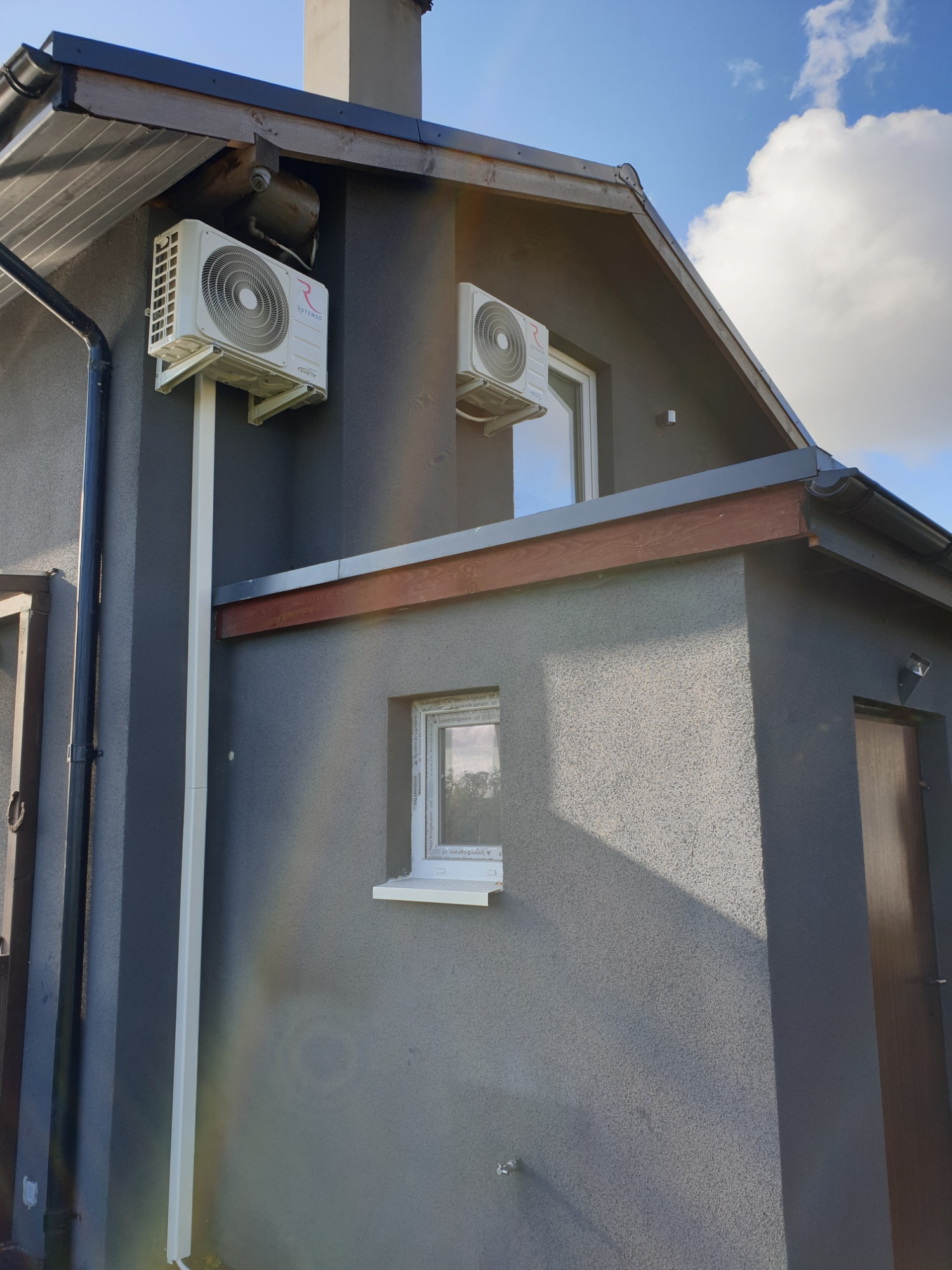 Dwa klimatyzatory marki Rotenso zamontowane na elewacji domu z szarą strukturą tynku, białe rury prowadzące w dół, błękitne niebo w tle.