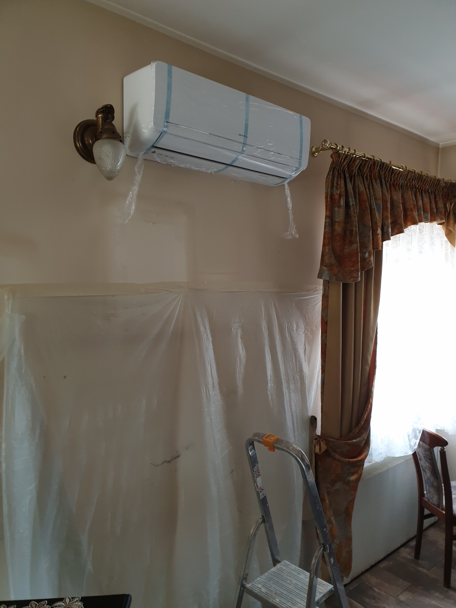 Nowo zainstalowany klimatyzator ścienny, jeszcze w folii ochronnej, zamontowany w pokoju z zasłoniętymi folią meblami i lampą ścienną w stylu retro, drabina pod ścianą.