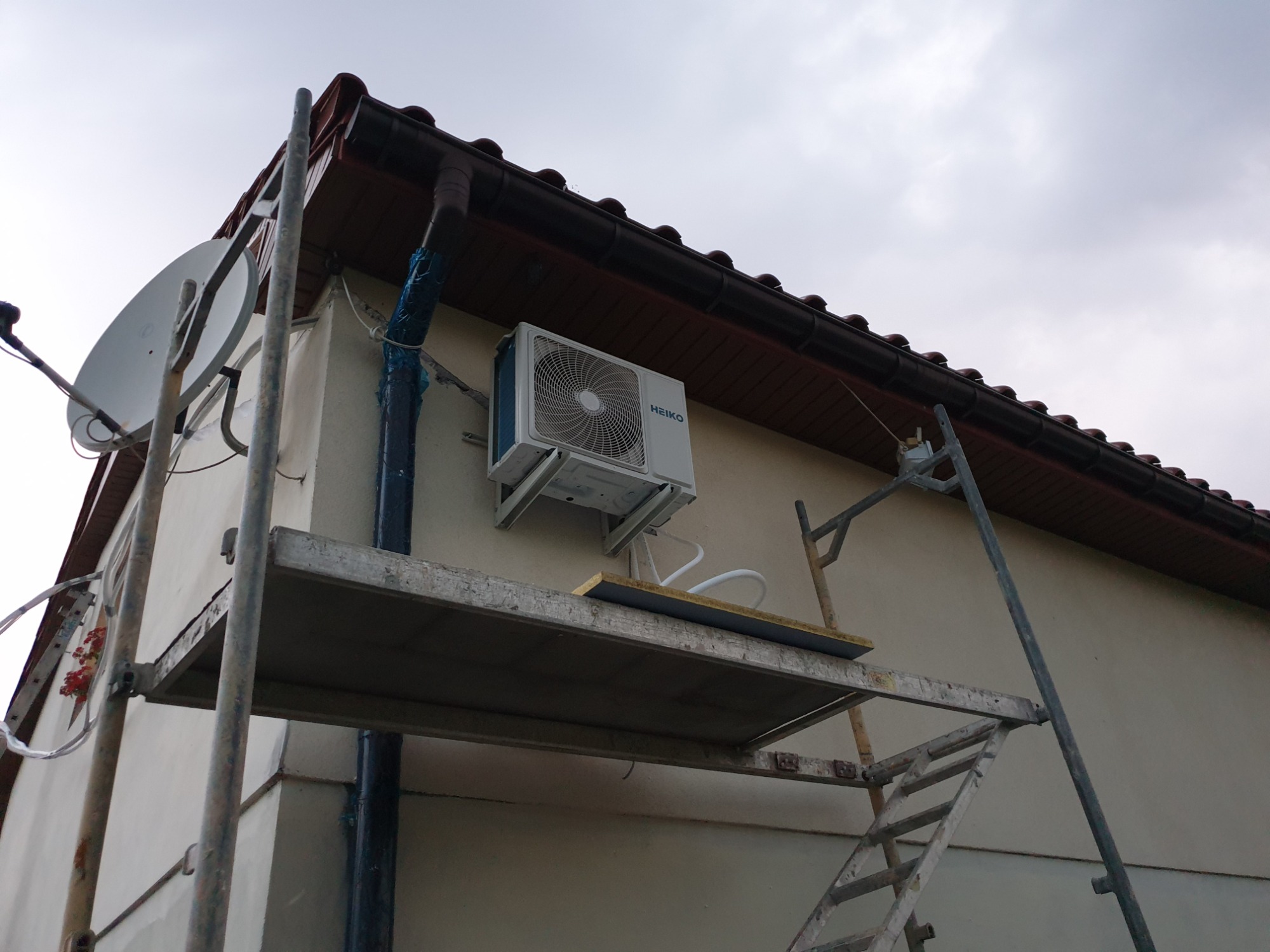 Zewnętrzna jednostka klimatyzacji marki Heiko zamontowana na elewacji domu, widoczna z rusztowania.