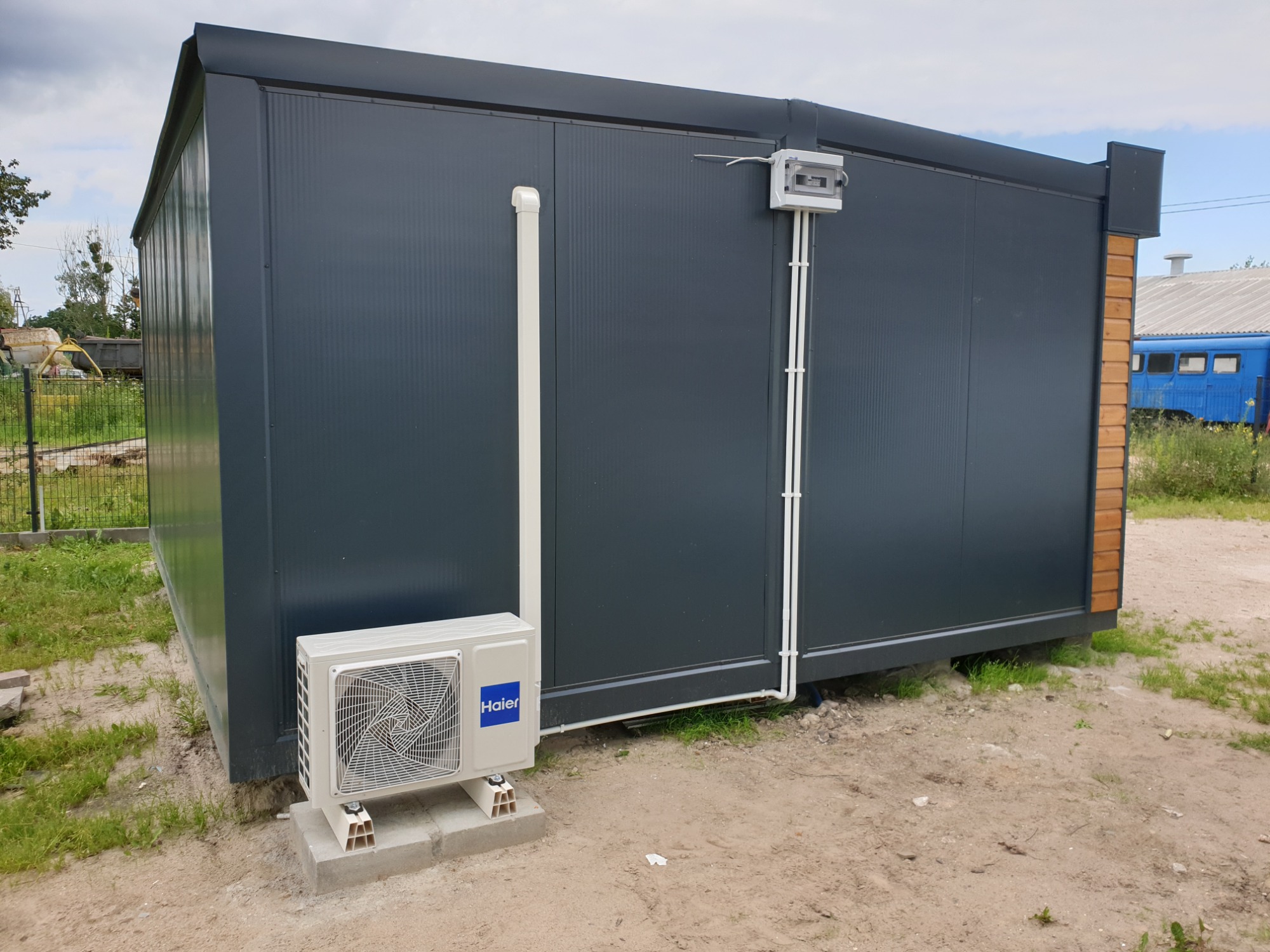 Zewnętrzna jednostka klimatyzacji Haier zamontowana na betonowych bloczkach przy szarym, metalowym kontenerze z widoczną instalacją elektryczną i rurami odprowadzającymi skropliny.