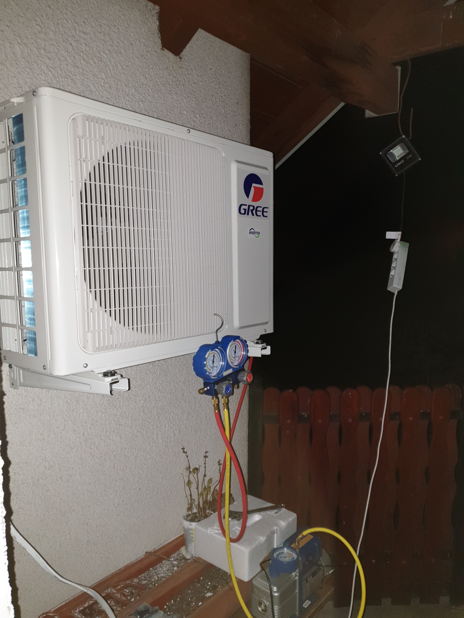 Biała jednostka zewnętrzna klimatyzacji marki Gree zamontowana na ścianie budynku, podłączona do manometrów serwisowych i pompy próżniowej podczas serwisu, widoczne przewody i narzędzia.