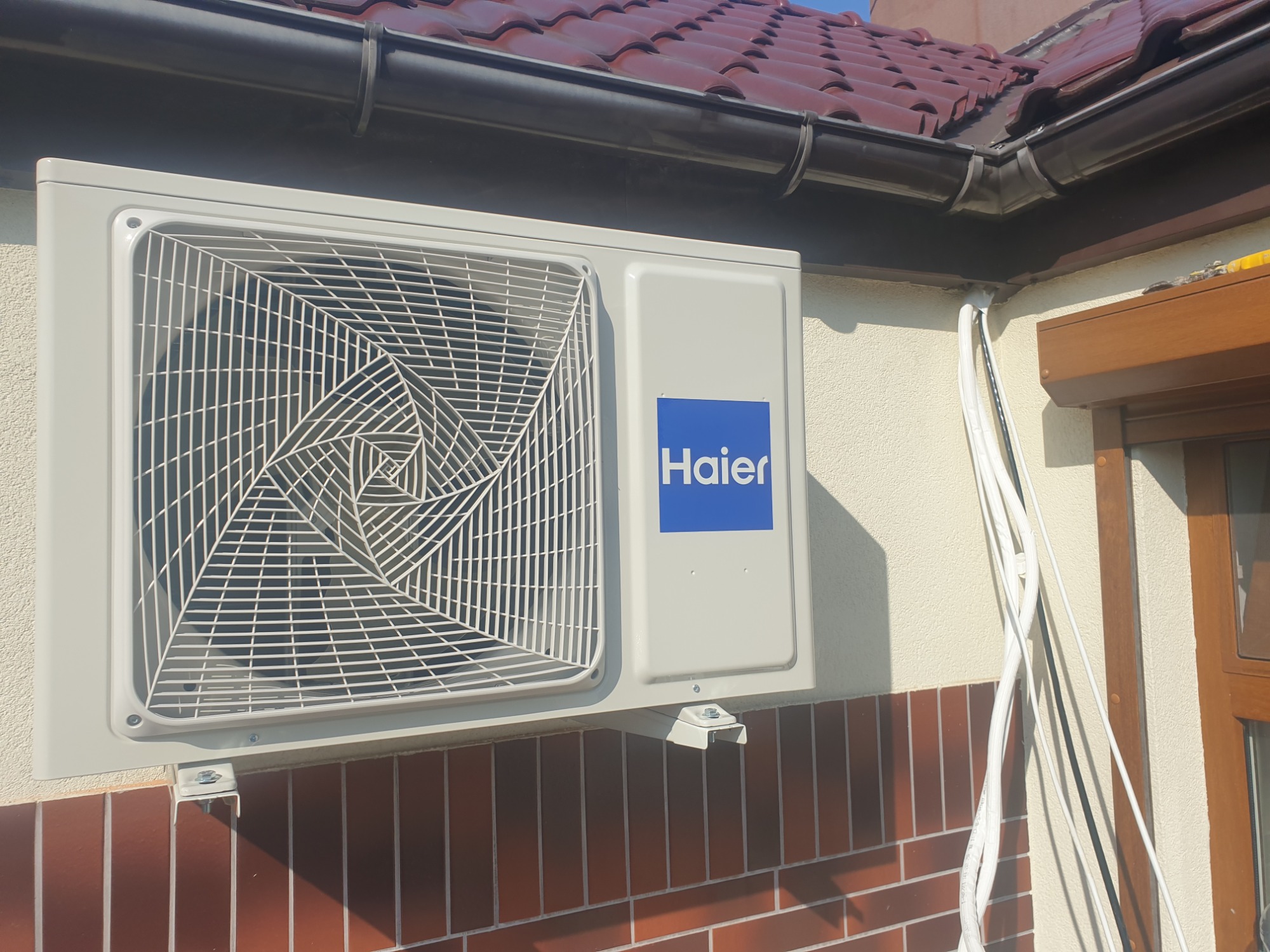 Zewnętrzna jednostka klimatyzacji Haier zamontowana na elewacji domu z ceglaną podmurówką, widoczne rury odprowadzające na zewnątrz budynku.