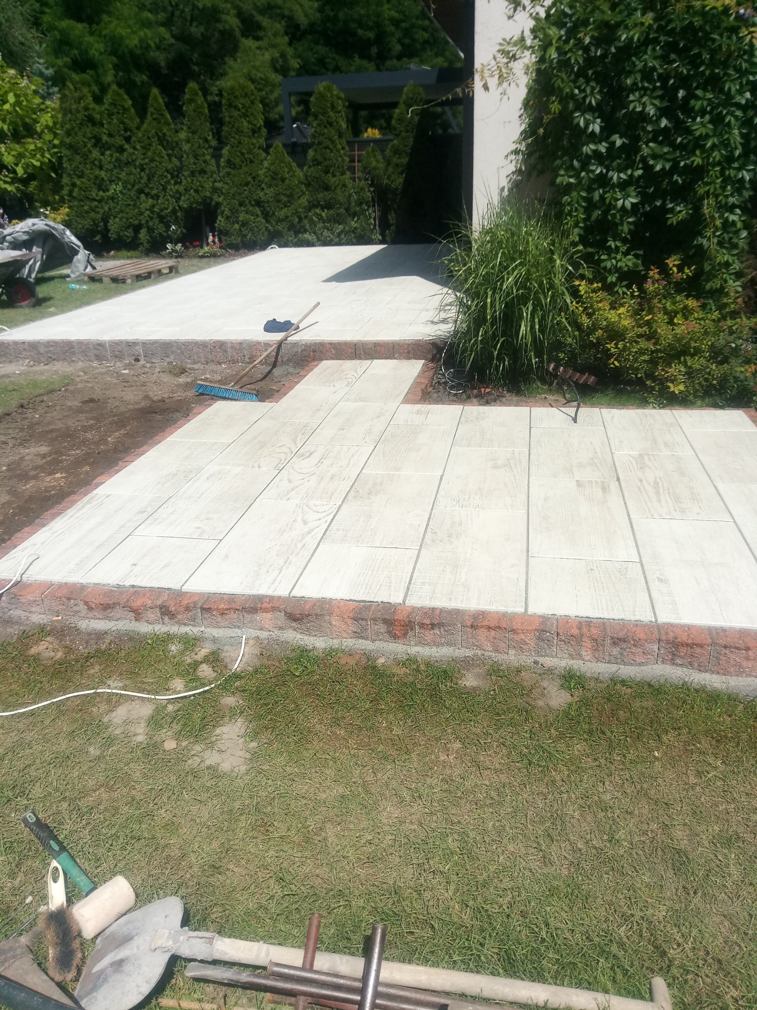 Nowo ułożony chodnik z jasnych płyt imitujących drewno, obramowany czerwoną cegłą, z widocznymi narzędziami brukarskimi i fragmentem ogrodu.