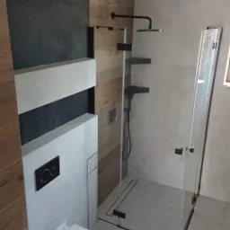 Nowoczesna łazienka z kabiną prysznicową typu walk-in z czarnymi detalami, podwieszana toaleta z czarnym przyciskiem spłukiwania, ściany wykończone płytkami imitującymi drewno i szarym kamieniem.