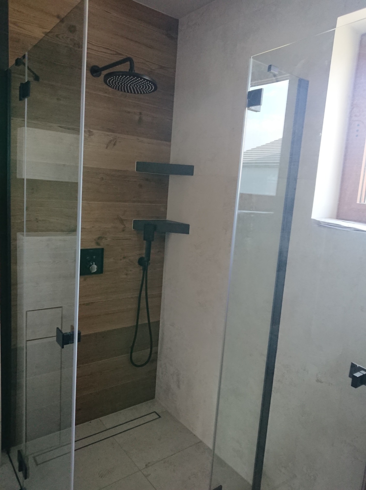 Nowoczesna kabina prysznicowa z deszczownicą, wykończona drewnopodobnymi płytkami i szarą ścianą, widoczny odpływ liniowy w podłodze.