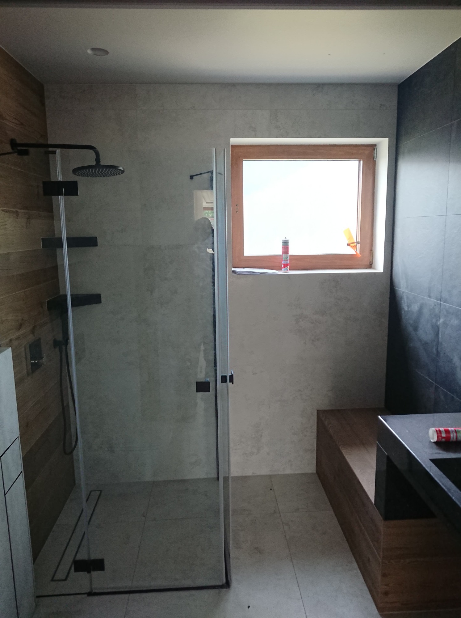 Nowoczesna łazienka z kabiną prysznicową typu walk-in, drewnianymi akcentami na ścianie i ławce, oraz oknem z tubą silikonu na parapecie.