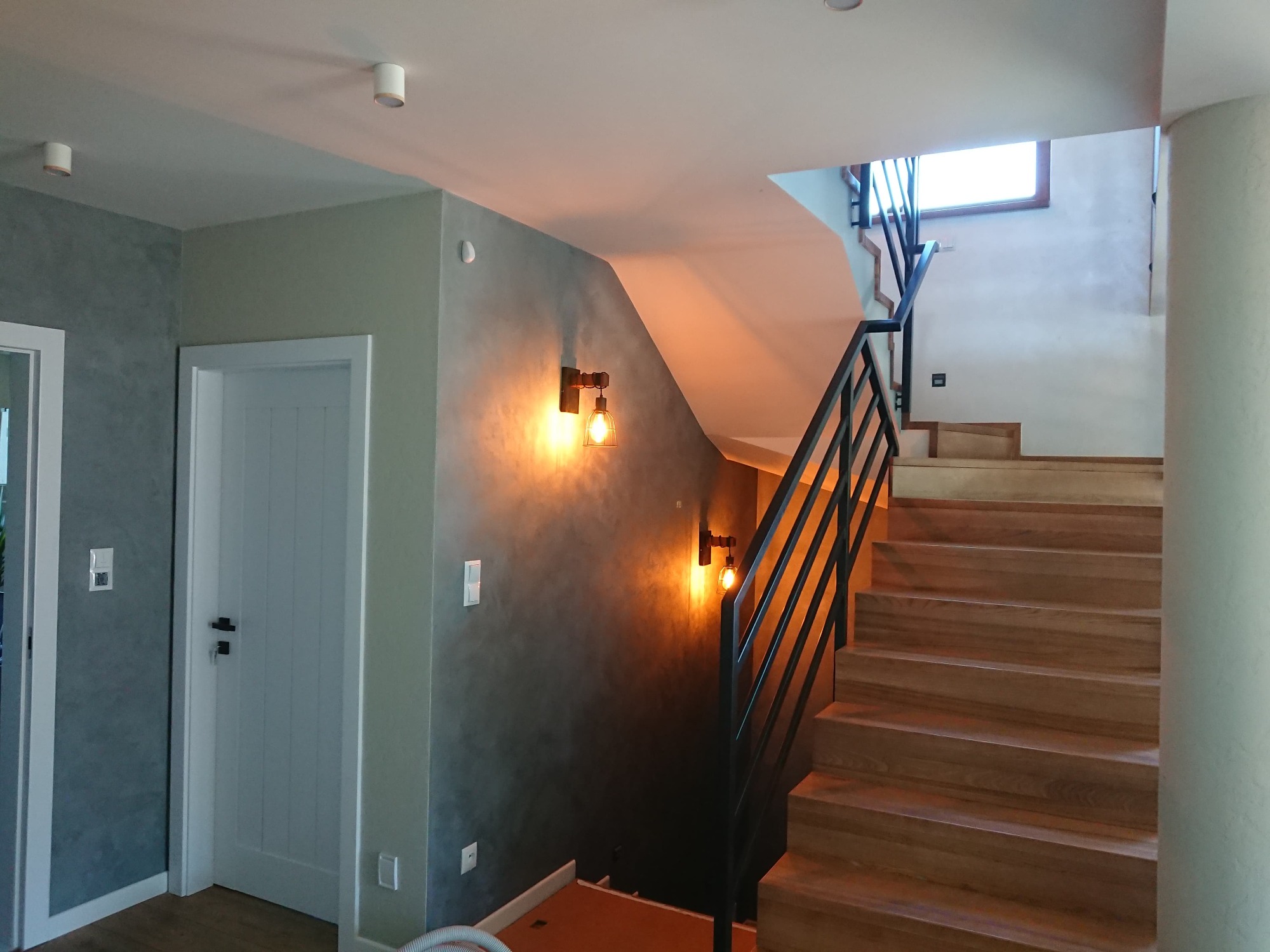 Klatka schodowa z drewnianymi schodami i czarną metalową balustradą, ściany w odcieniach szarości z dekoracyjnym wykończeniem, oświetlone dwoma kinkietami w stylu industrialnym.