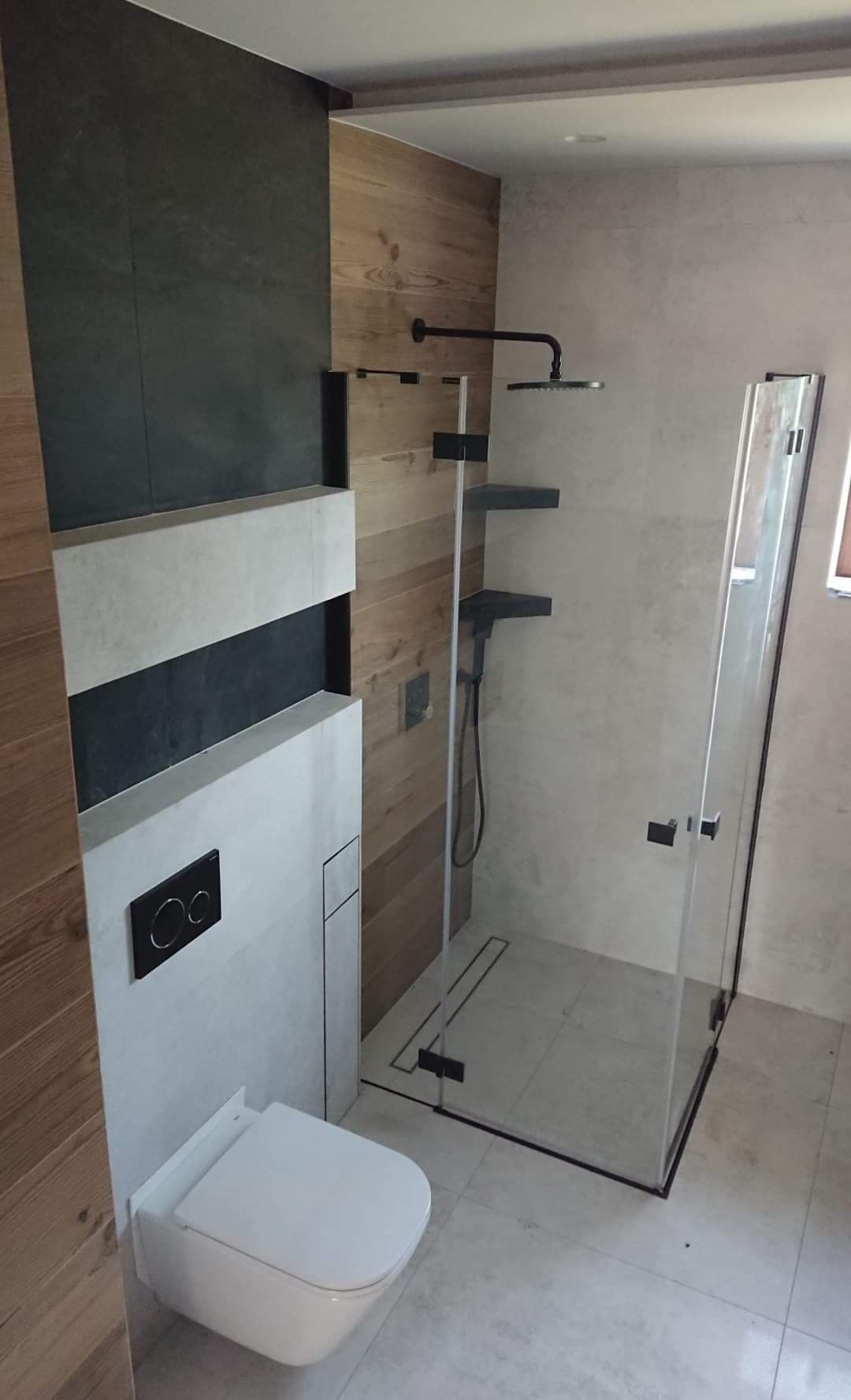 Nowoczesna łazienka z kabiną prysznicową typu walk-in z czarnymi detalami, podwieszana toaleta z czarnym przyciskiem spłukiwania, ściany wykończone płytkami imitującymi drewno i szarym kamieniem.