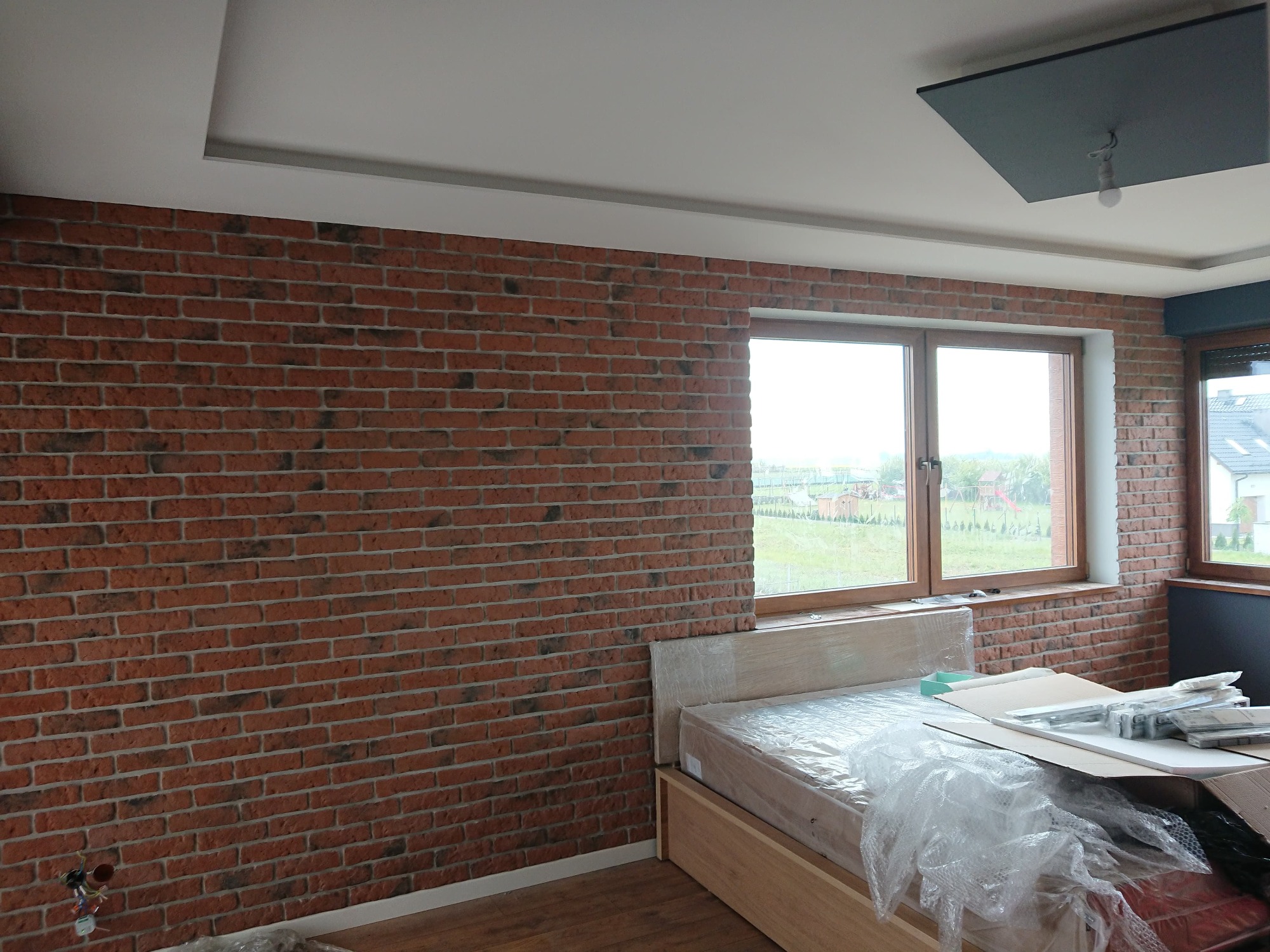 Pokój w trakcie wykańczania: ściana z cegły, okna z widokiem na zieleń, łóżko zabezpieczone folią, kartony i materiały budowlane.