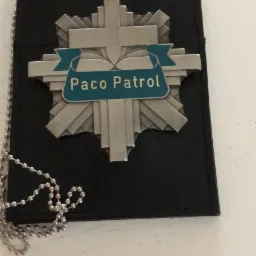 Metalowa odznaka 'Paco Patrol' na czarnym skórzanym etui, z łańcuszkiem kulkowym, leżąca na białym tle w Gliwicach.