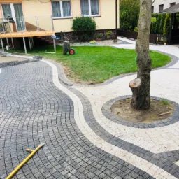 Kaskadowy wzór szarej i białej kostki brukowej tworzy falisty chodnik przed domem z drewnianym tarasem, w tle widoczny trawnik i altana.