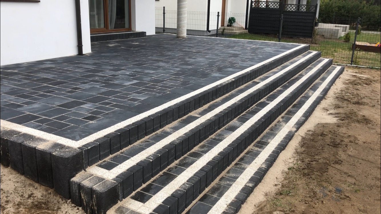 Ułożony taras z ciemnej kostki brukowej z jasnymi fugami i schodami, widok z boku na tle domu i ogrodu.