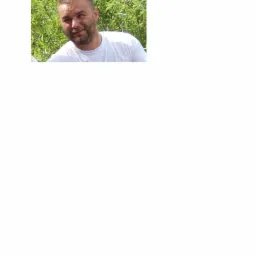 Portret mężczyzny w białej koszulce na tle zielonych liści, uśmiechający się w naturalnym oświetleniu.