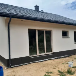 Nowoczesny dom jednorodzinny z jasną elewacją i ciemną podmurówką, widoczne okna z jasnymi ramami, czarny dach z blachodachówki i rynny, teren wokół domu w trakcie prac wykończeniowych, widoczne...