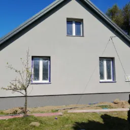 Świeżo otynkowana elewacja domu jednorodzinnego w odcieniach szarości, z zabezpieczonymi oknami folią, widoczny fragment ogrodu i małe drzewko.