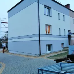 Dwukondygnacyjny budynek po częściowym ociepleniu, z widocznym rusztowaniem, beczką Orlen Oil i pracownikami w trakcie prac wykończeniowych.