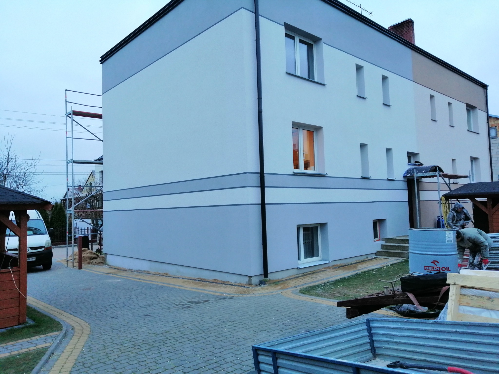 Dwukondygnacyjny budynek po częściowym ociepleniu, z widocznym rusztowaniem, beczką Orlen Oil i pracownikami w trakcie prac wykończeniowych.