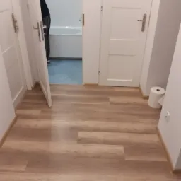 Widok na korytarz z nowo położonymi panelami podłogowymi w odcieniach drewna, z widocznymi białymi drzwiami do łazienki z wanną i osobą w środku oraz rolką papieru toaletowego przy ścianie.