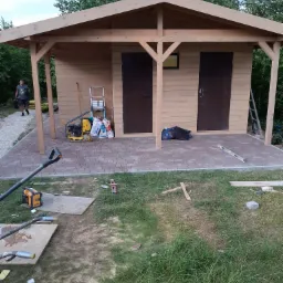 Drewniana altana z dwoma wejściami, wyłożona kostką brukową, w trakcie budowy, widoczne narzędzia i materiały budowlane, na trawie leżą resztki materiałów.