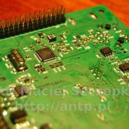 Fabryka Aparatury Nietypowej Maciej Szczepka - Zielona płytka drukowana z elementami elektronicznymi, widoczne układy scalone, kondensatory i złącza pinowe, oświetlona ciepłym światłem, z naniesionym adresem strony internetowej na powierzchni.