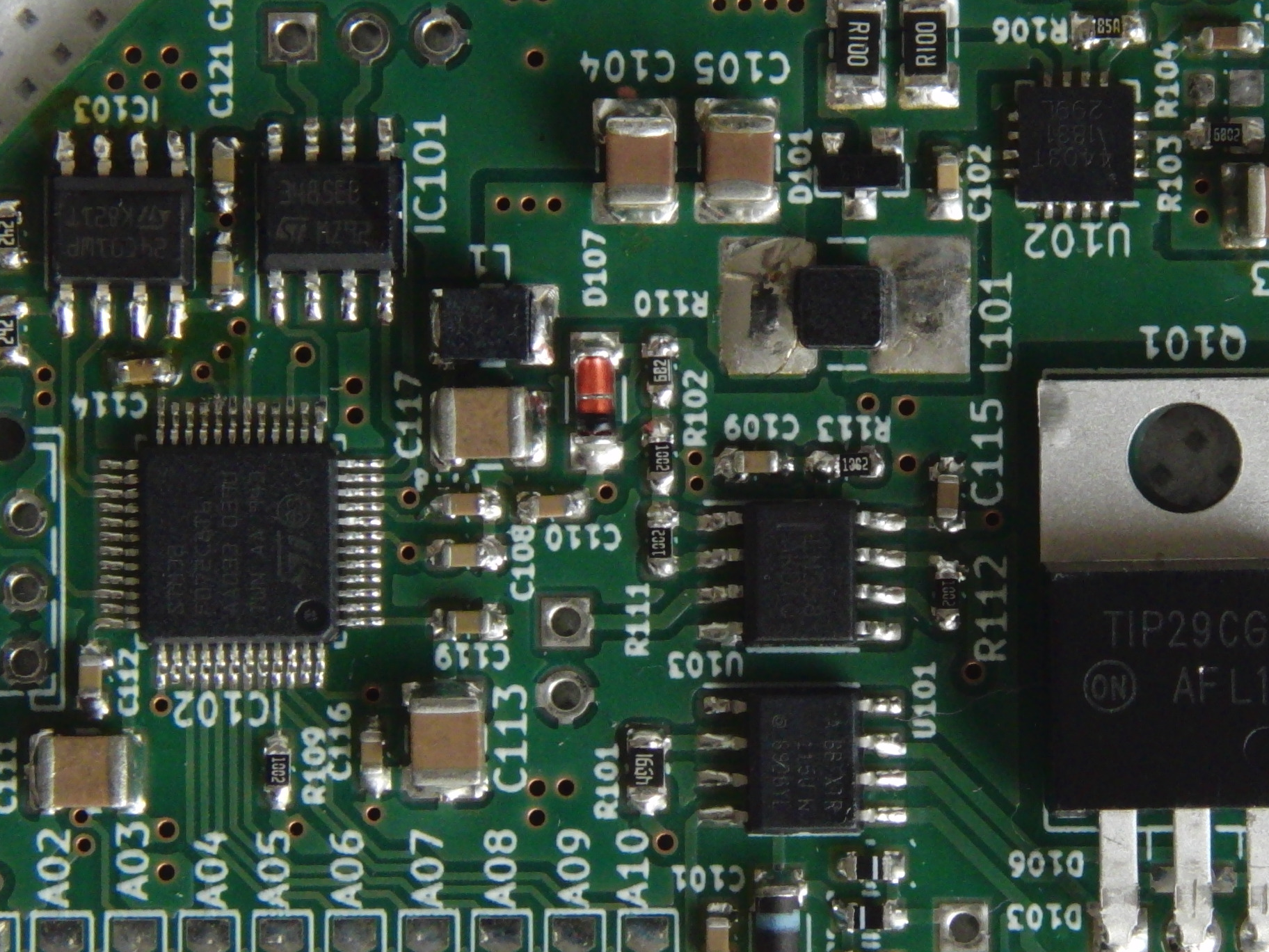 Zbliżenie na zieloną płytę drukowaną z licznymi elementami elektronicznymi, w tym układy scalone, rezystory i kondensatory, z widocznymi oznaczeniami i numerami komponentów.