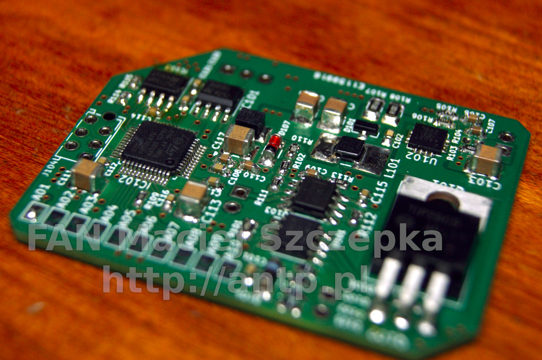 Zielona płytka drukowana z elementami elektronicznymi, widoczne układy scalone, rezystory i kondensatory, z bliska na drewnianym tle.