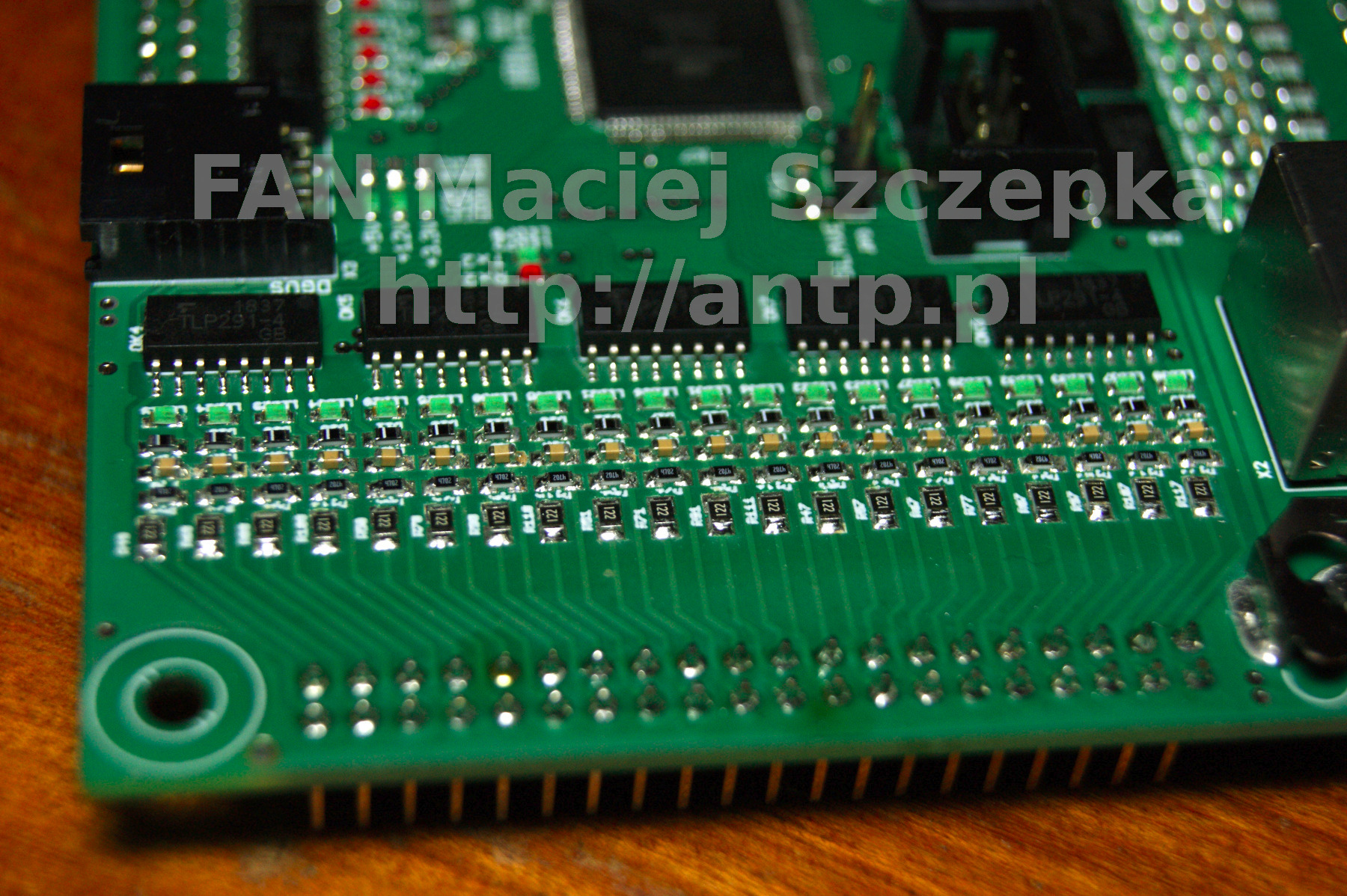 Fragment zielonej płytki drukowanej z rzędami komponentów elektronicznych, widoczne układy scalone i ścieżki, nazwa firmy FAN Maciej Szczepka i adres strony http://antp.pl naniesione na płytkę.
