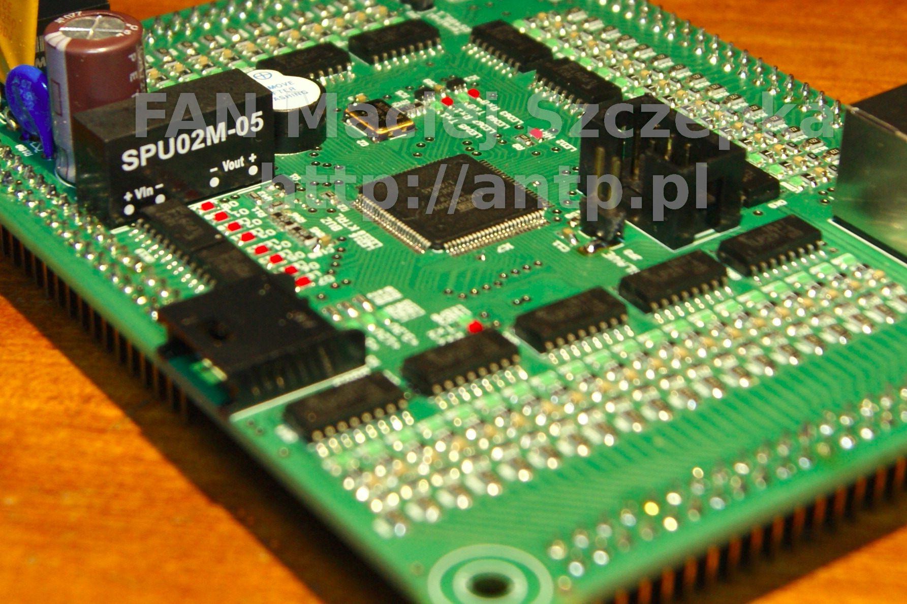 Zielona płytka drukowana z elementami elektronicznymi, widoczne układy scalone, kondensatory i złącza, leżąca na drewnianej powierzchni.