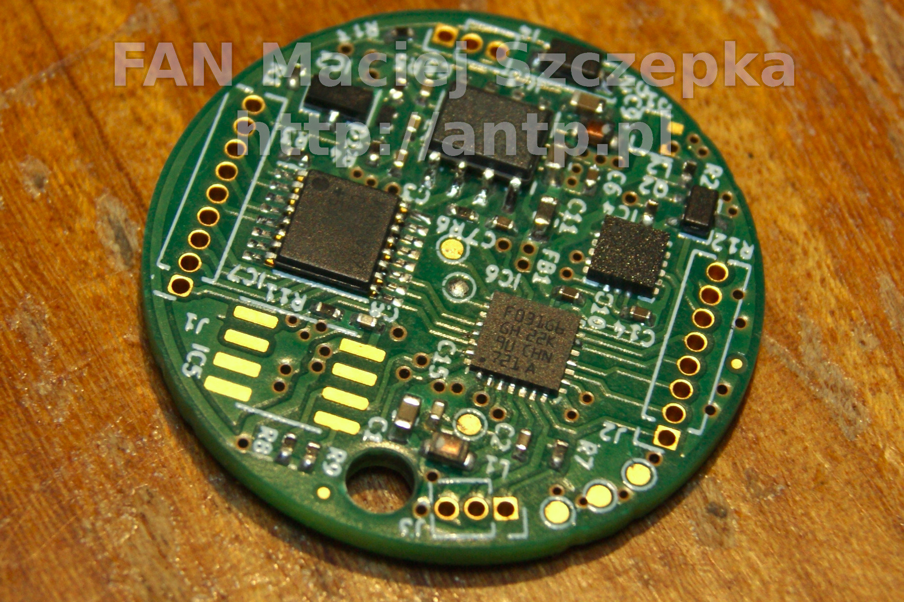 Okrągła płytka drukowana w kolorze zielonym z widocznymi elementami elektronicznymi, takimi jak układy scalone, rezystory i kondensatory, umieszczona na drewnianej powierzchni.