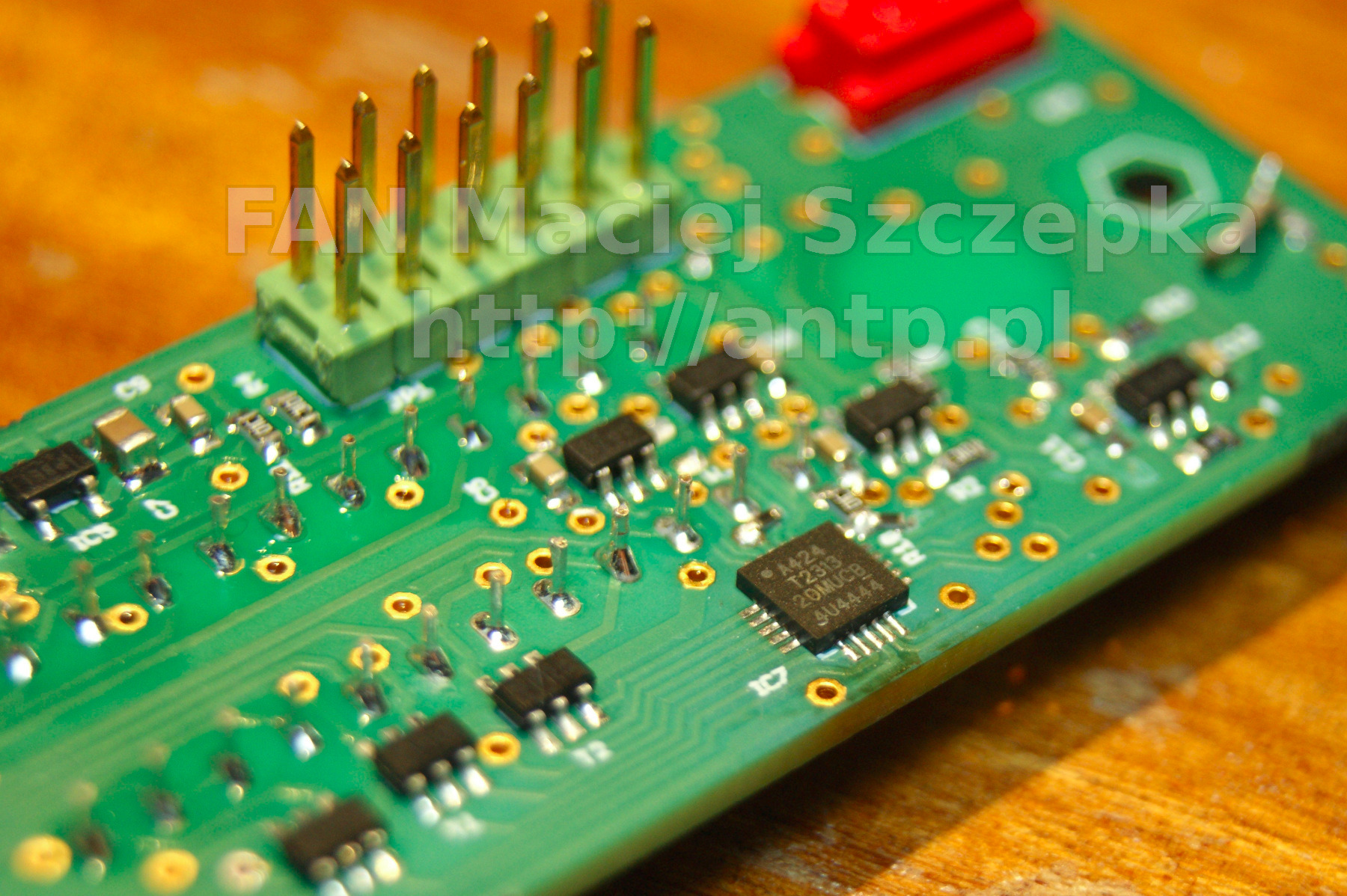 Zbliżenie na zieloną płytkę drukowaną z elementami elektronicznymi, w tym układ scalony z napisem 'A424 T2313 20MUCB 4U4444' i złączem pinowym z pozłacanymi pinami, leżącą na drewnianej powierzchni.