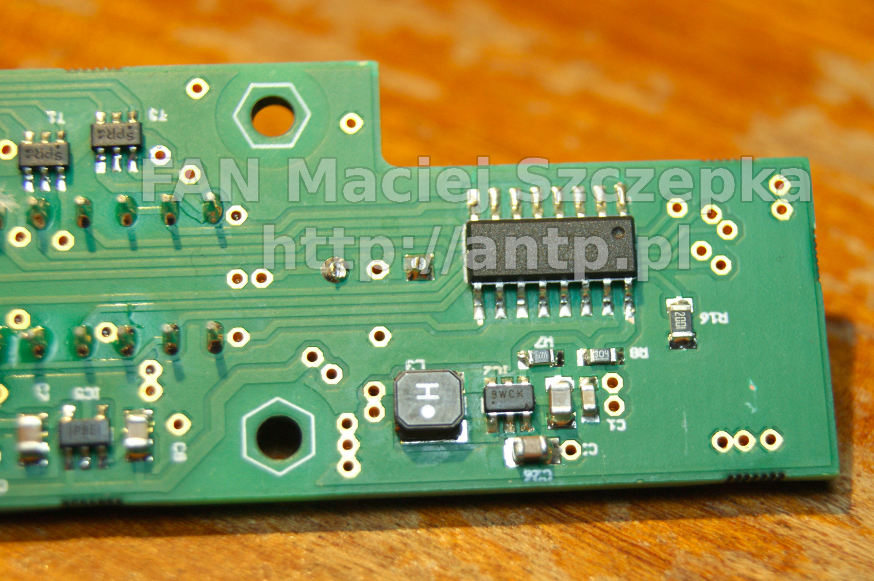 Fragment zielonej płytki drukowanej z elementami elektronicznymi, widoczne układy scalone, rezystory i kondensatory, z bliska, na drewnianym podłożu.