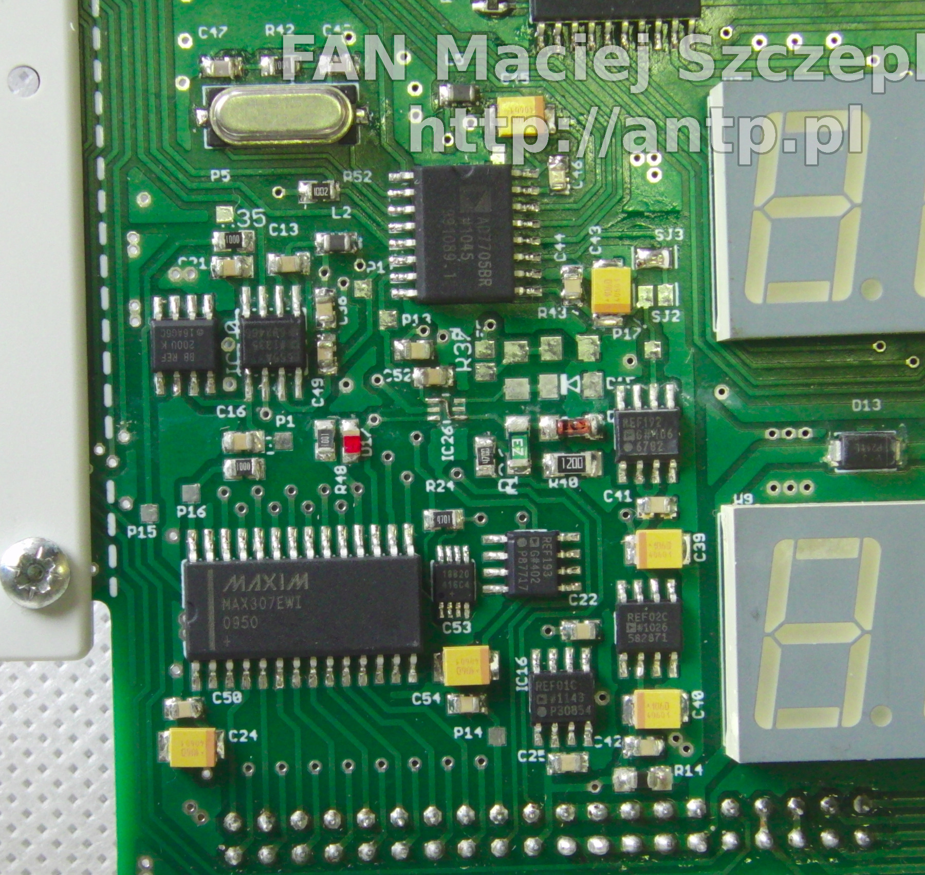 Zbliżenie na fragment zielonej płytki drukowanej z licznymi elementami elektronicznymi, w tym układy scalone (np. MAXIM MAX307EWI), kondensatory, rezystory i wyświetlacze siedmiosegmentowe.