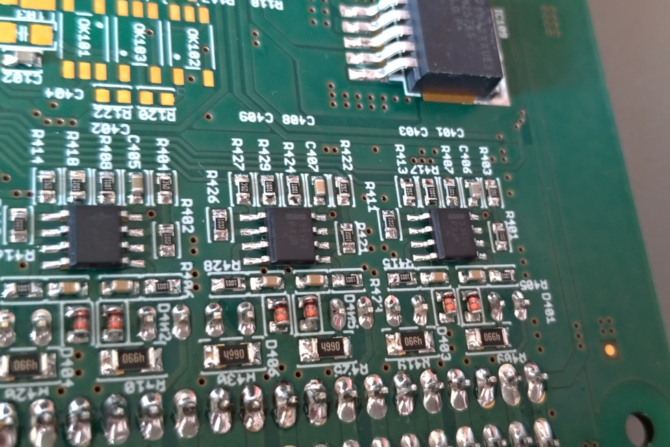 Szczegółowe ujęcie fragmentu zielonej płyty głównej z przylutowanymi elementami elektronicznymi, widoczne rezystory, kondensatory i układy scalone z precyzyjnymi oznaczeniami.