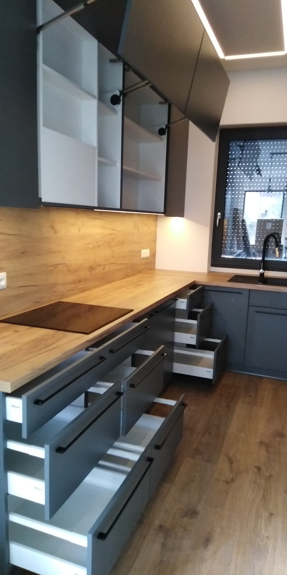 Szare szafki kuchenne z drewnianym blatem, otwarte szuflady z systemem Blum, górne szafki podnoszone do góry, oświetlenie LED podszafkowe.