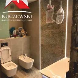 Wykończona łazienka z wiszącą toaletą i bidetem, umywalką z podświetleniem LED, dekoracyjnymi płytkami i lampami w folii ochronnej, z widocznym logo firmy remontowej.