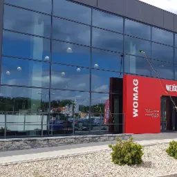 Czyszczenie szklanej fasady budynku Womag za pomocą lancy teleskopowej z osmozą, widoczne odbicie nieba i otoczenia w oknach.