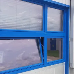 Błękitne okno z ramą z PCV, jedno skrzydło uchylone, widoczne żaluzje w górnej części i odbicie chmur w szybach.