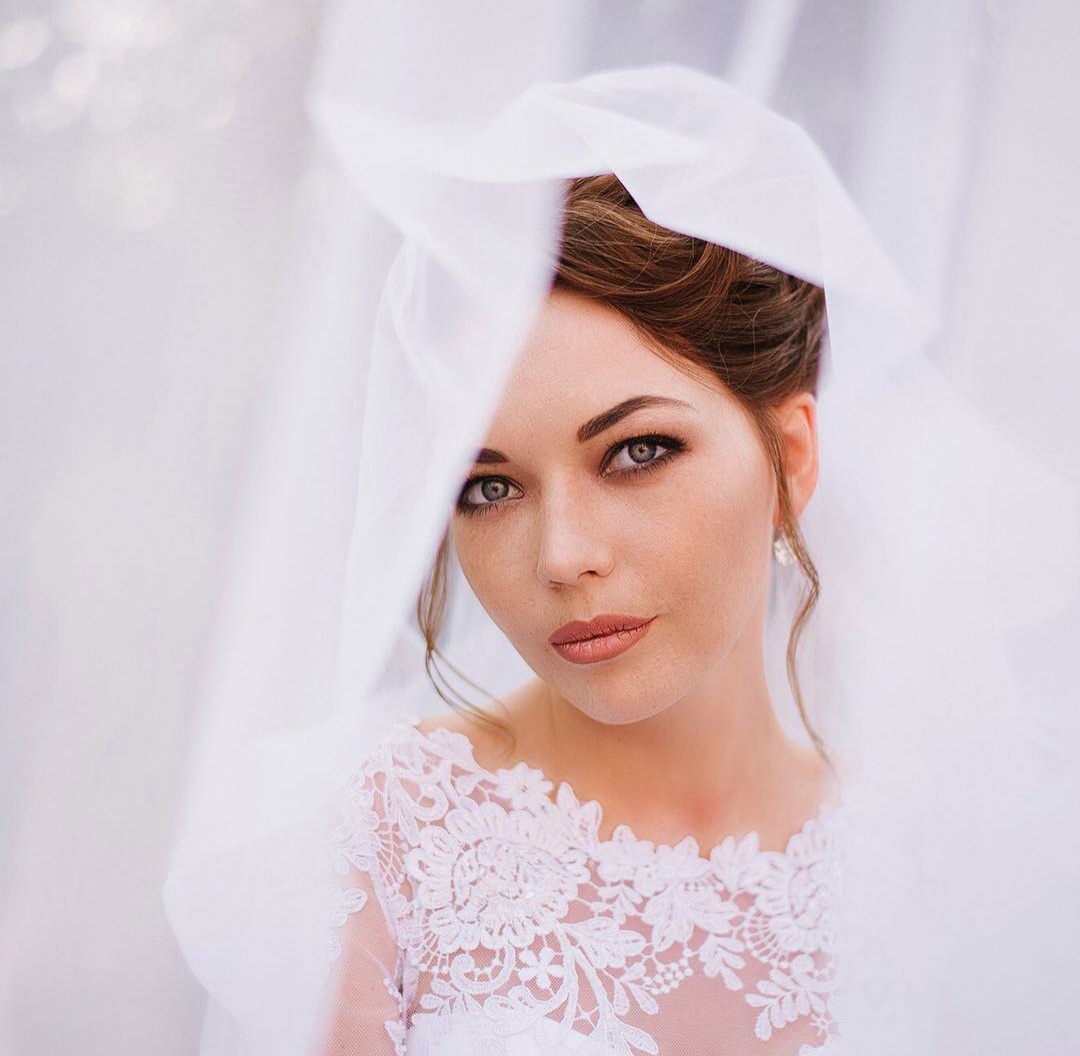 Portret panny młodej z delikatnym makijażem oczu i ust, częściowo zasłoniętej przez biały welon z koronkowym wykończeniem dekoltu sukni ślubnej.