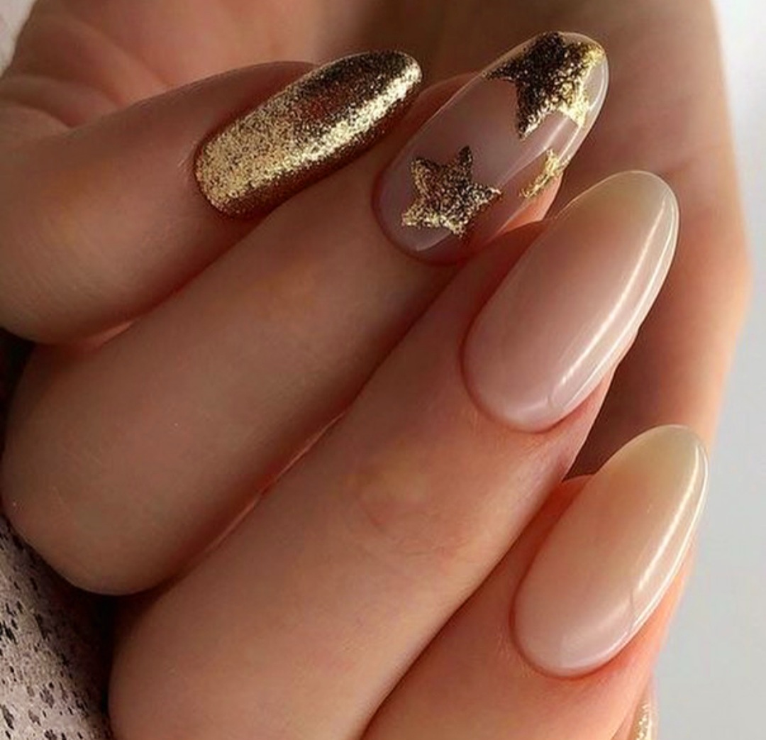 Dłoń z owalnymi paznokciami: jeden pokryty złotym brokatem, drugi z naklejonymi złotymi gwiazdkami na przezroczystym tle, pozostałe w odcieniu nude.