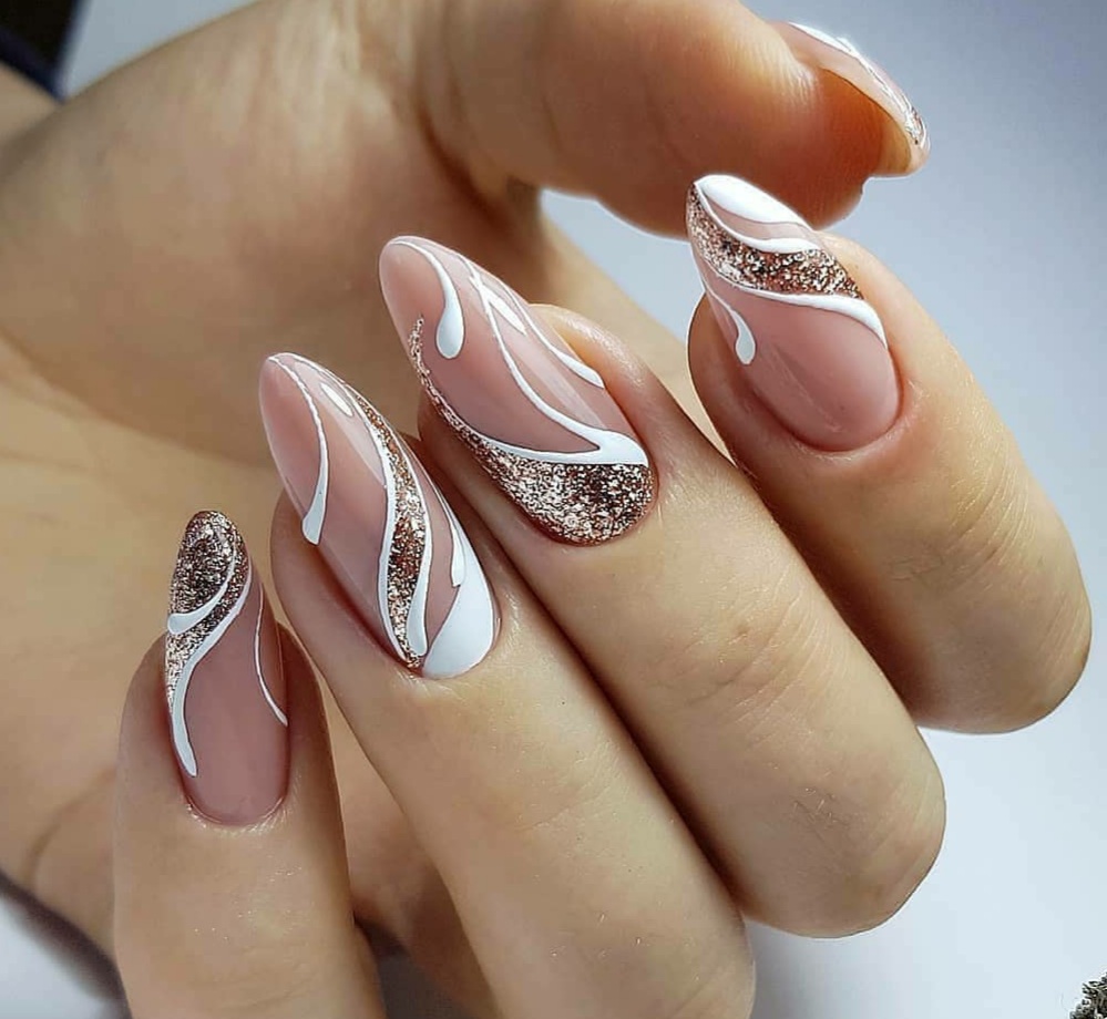 Dłoń z beżowymi paznokciami o migdałowym kształcie, zdobionymi białymi, abstrakcyjnymi liniami i brokatem w kolorze różowego złota.