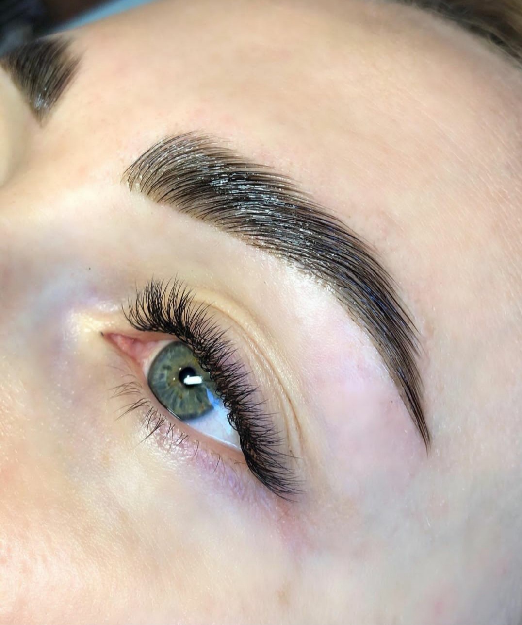 Zbliżenie na oko z długimi, czarnymi rzęsami i wystylizowaną, błyszczącą brwią o ciemnobrązowym odcieniu, ukazujące efekt laminacji.