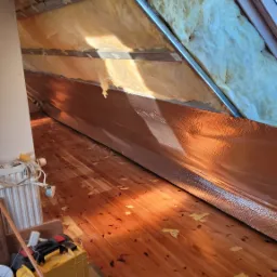 Poddasze w trakcie ocieplania, widoczne wełna mineralna między krokwiami, folia aluminiowa, drewniana podłoga z resztkami materiału izolacyjnego, narzędzia pracy i okno dachowe.