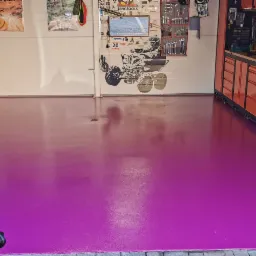 Wnętrze garażu z intensywnie fioletową, błyszczącą posadzką żywiczną, widoczne narzędzia na ścianach i szafkach warsztatowych.