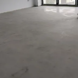 Szara, gładka posadzka w jasnym pomieszczeniu z dużymi oknami, widoczny fragment ściany z gniazdkiem elektrycznym.
