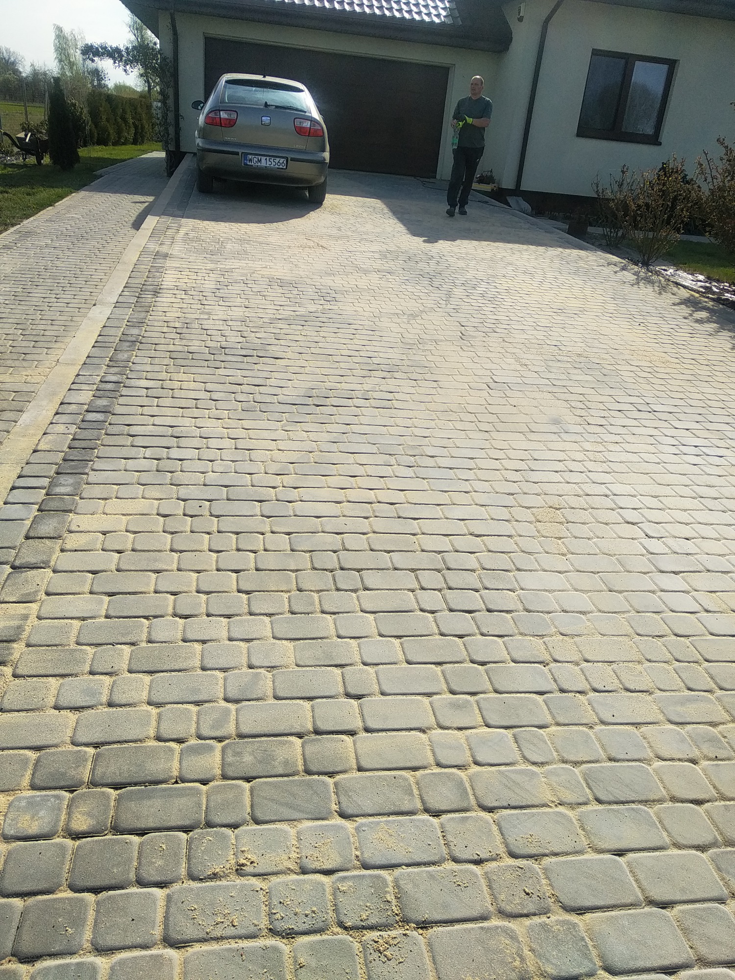 Ułożona szara kostka brukowa na podjeździe przed domem z beżową elewacją i brązową bramą garażową; widoczny samochód osobowy i osoba stojąca z boku.