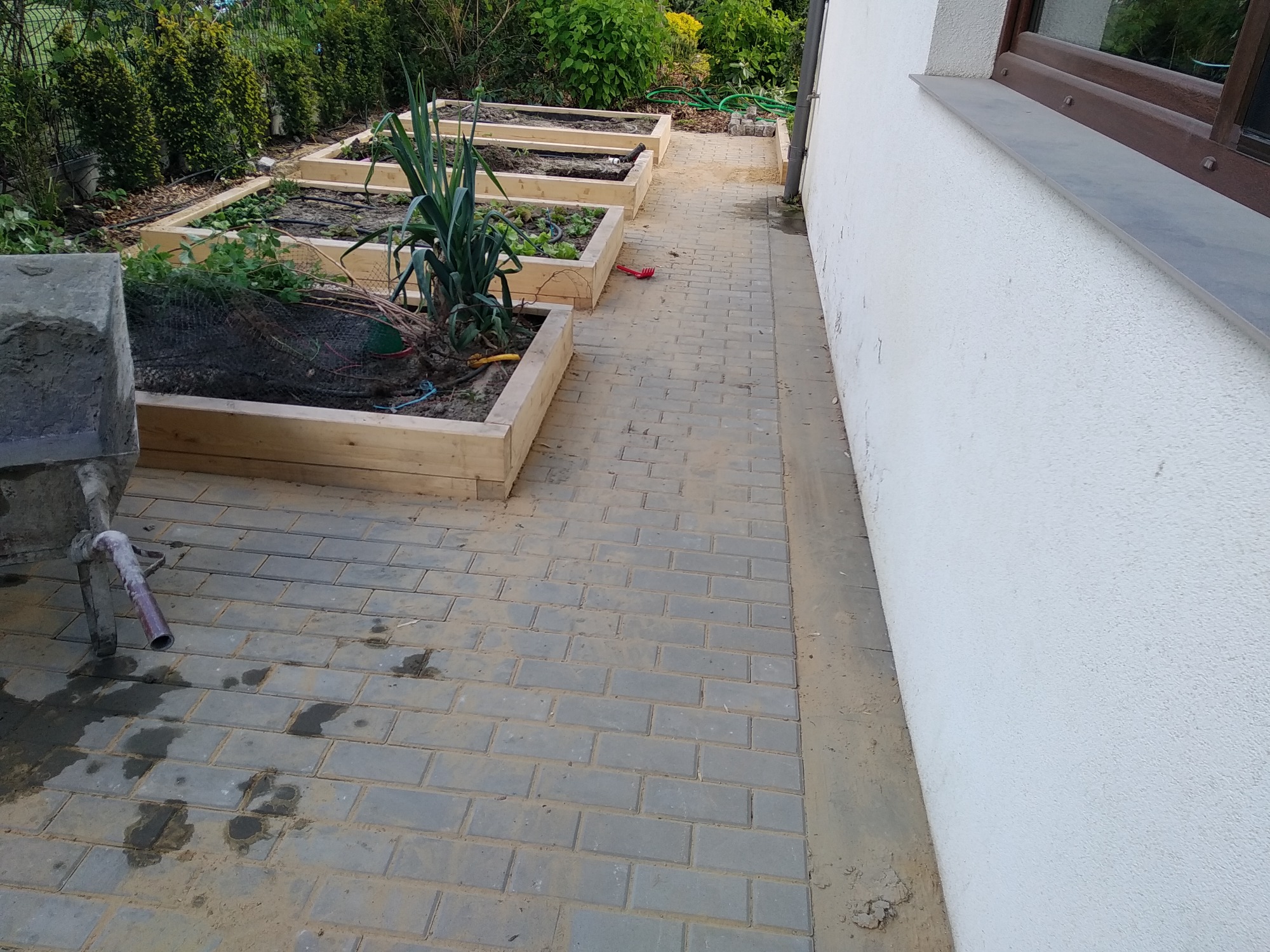 Szara kostka brukowa ułożona wzdłuż białej ściany budynku, z drewnianymi podwyższonymi grządkami warzywnymi w tle, wózek budowlany z resztkami zaprawy po lewej stronie.