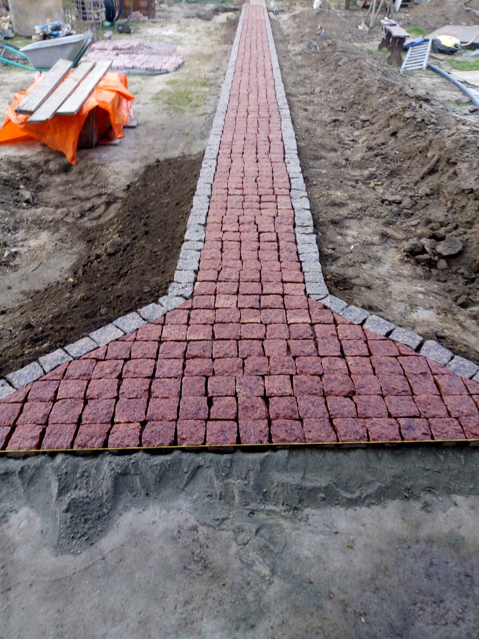Fragment ścieżki z czerwonej kostki granitowej z szarą obwódką, w trakcie układania, widoczne przygotowane podłoże i narzędzia w tle.