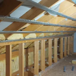 Poddasze w trakcie ocieplania: widoczne drewniane krokwie, wełna mineralna, metalowe profile oraz fragment murowanego komina i okno.