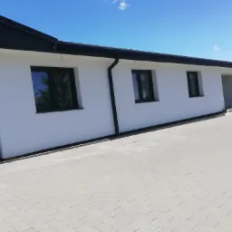 Biały, parterowy budynek z ciemnymi oknami i dachem, widziany z perspektywy chodnika z kostki brukowej w słoneczny dzień.